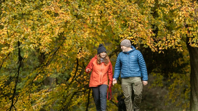 Woman in red waterproof jacket and man in blue puffer jacket walking with a small black dog on a woodland path